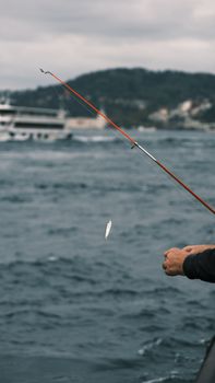 A person fishing by the sea with a visible fishing rod and distant ferry.
