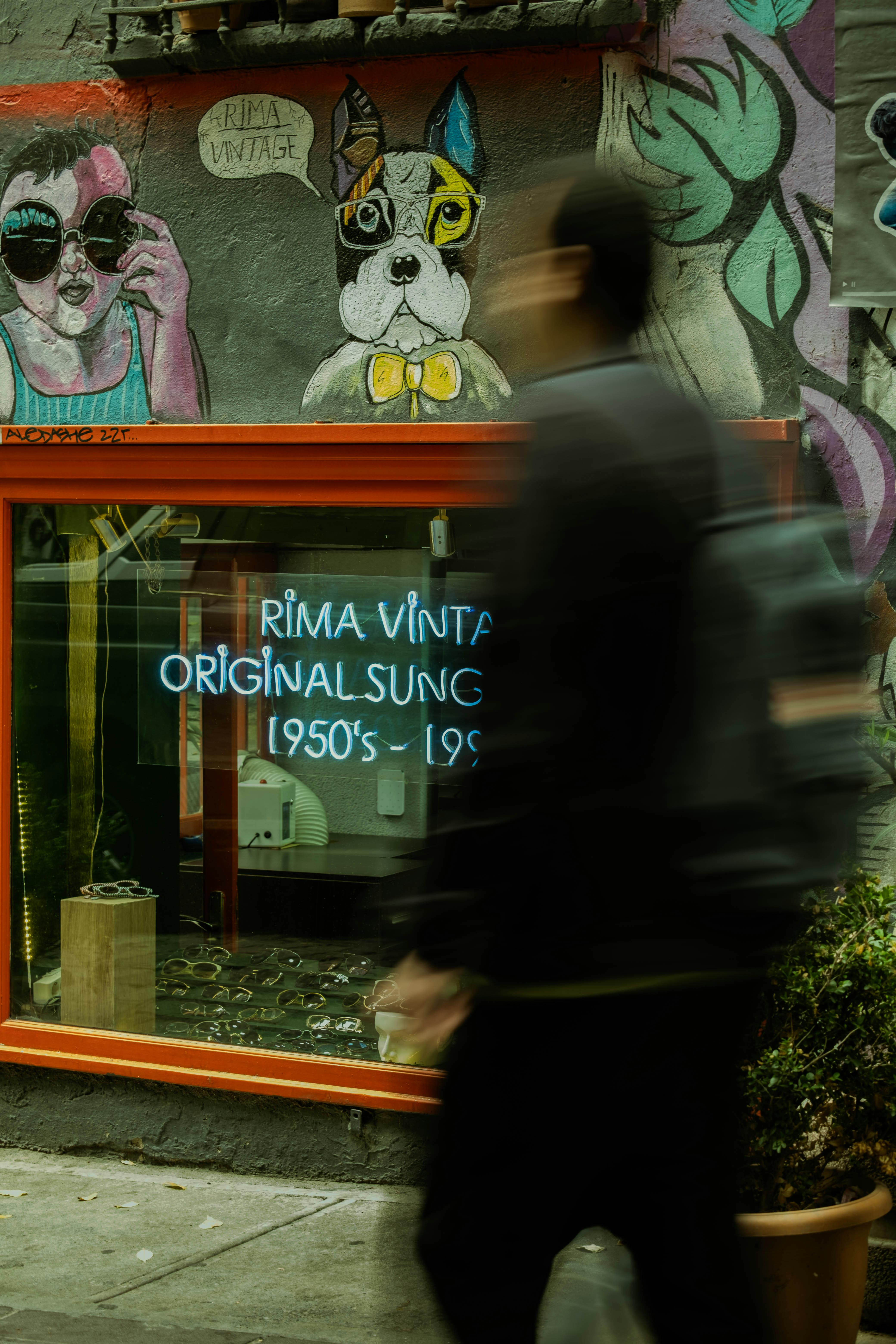 Blurred person passing a vintage sunglasses store with colorful murals.