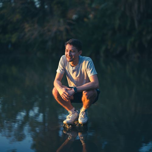 Free Man squatting near a calm lake at sunset, enjoying nature's serenity. Stock Photo