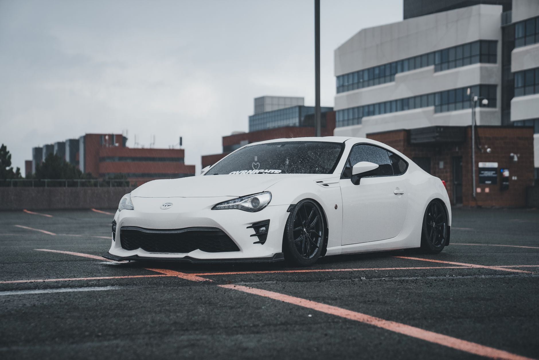 White sports car parked in urban area with overcast sky, modern architecture backdrop.