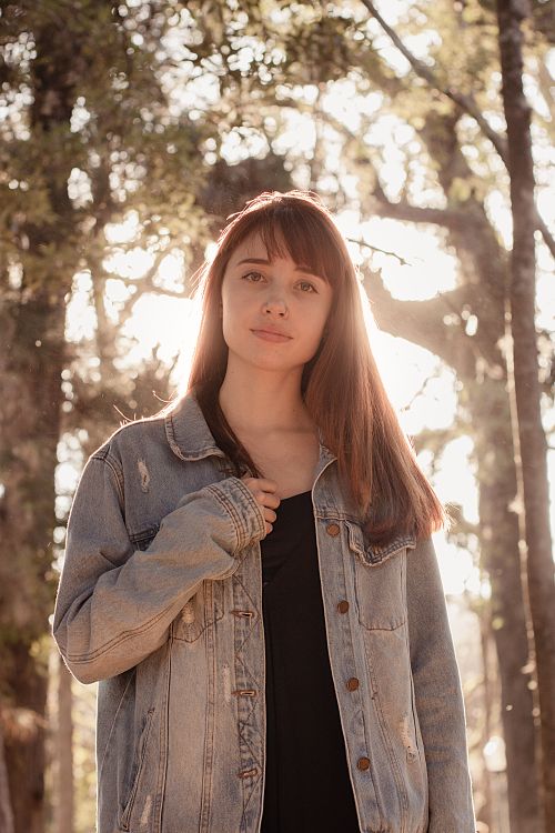 Free Young woman with a serene expression in a sunlit forest, wearing a denim jacket. Stock Photo