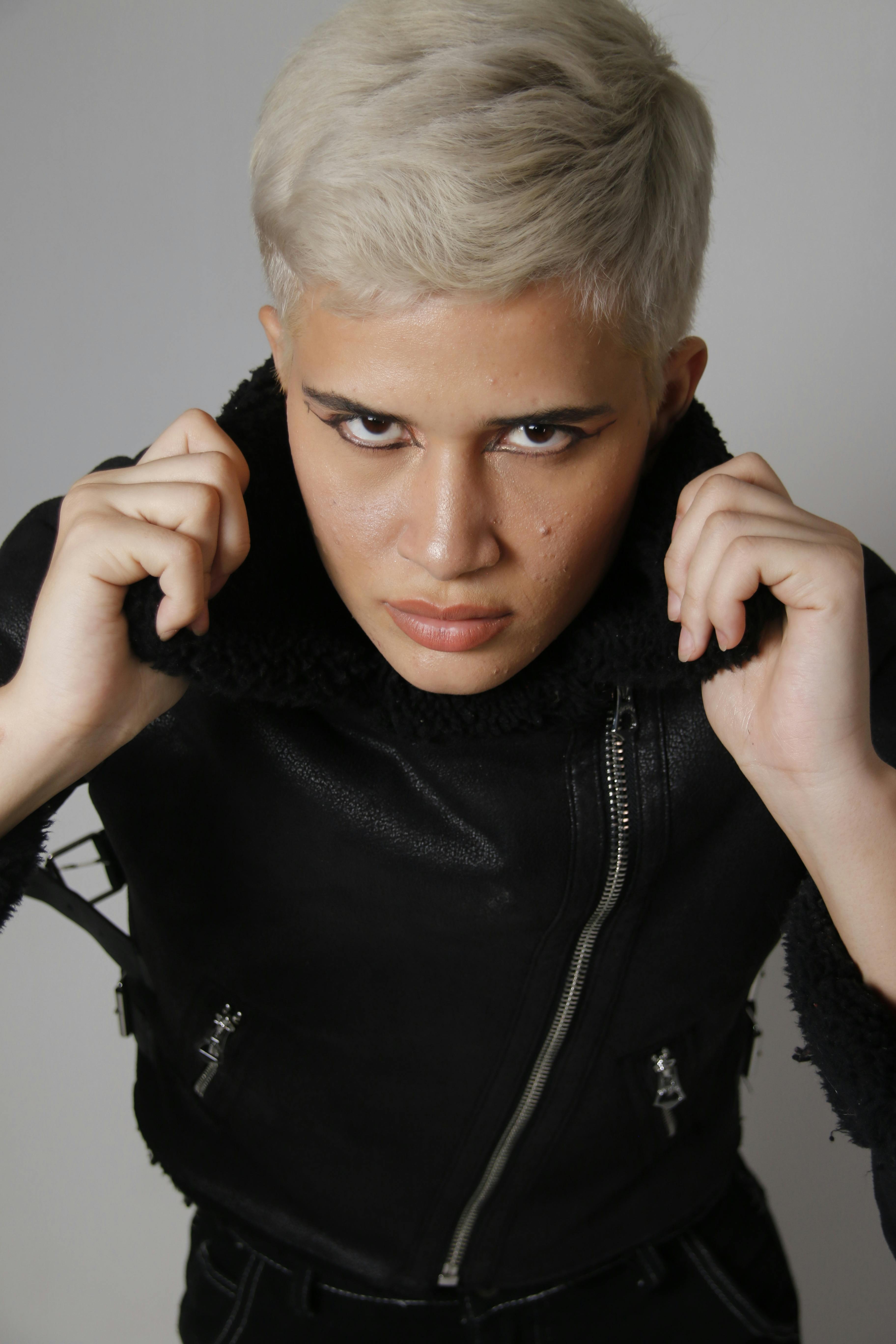 Confident young adult with blonde hair poses in stylish leather jacket, exuding edgy and modern fashion.