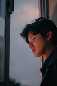 Young man with artistic makeup gazes out window at dusk, creating a mysterious mood.