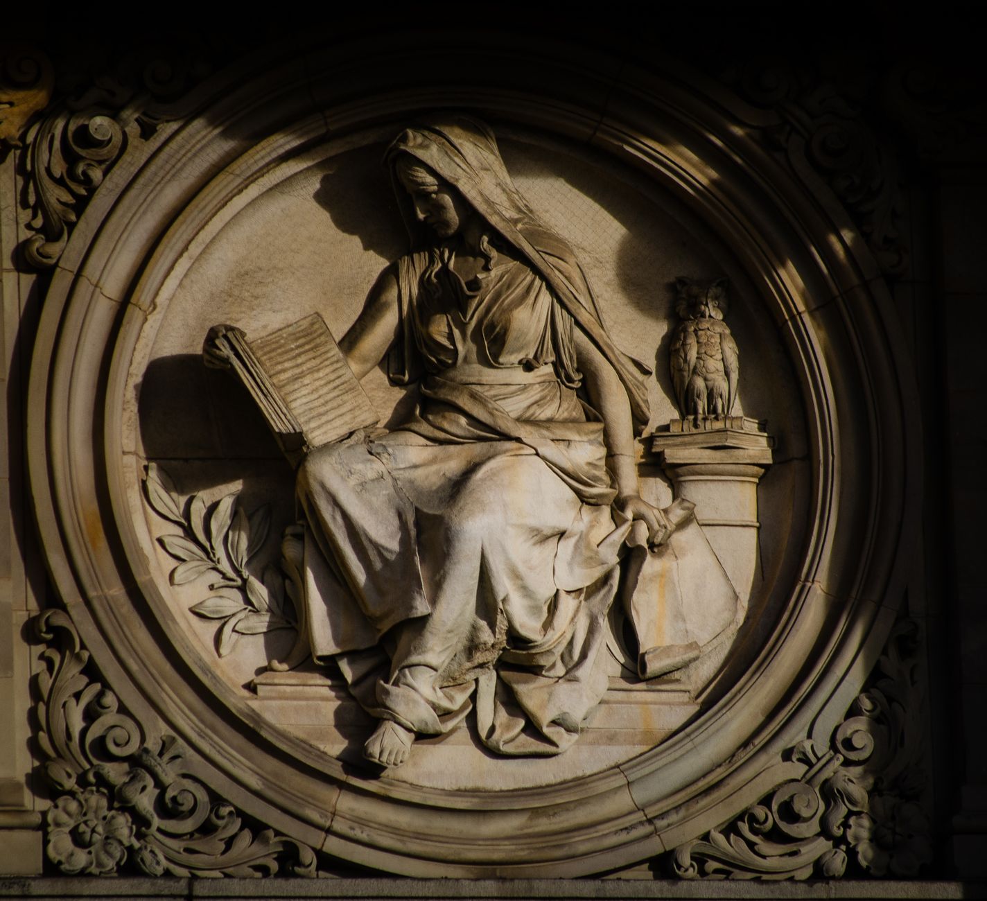 Elegant stone carving of a robed woman reading, accompanied by an owl, on an ornate roundel.
