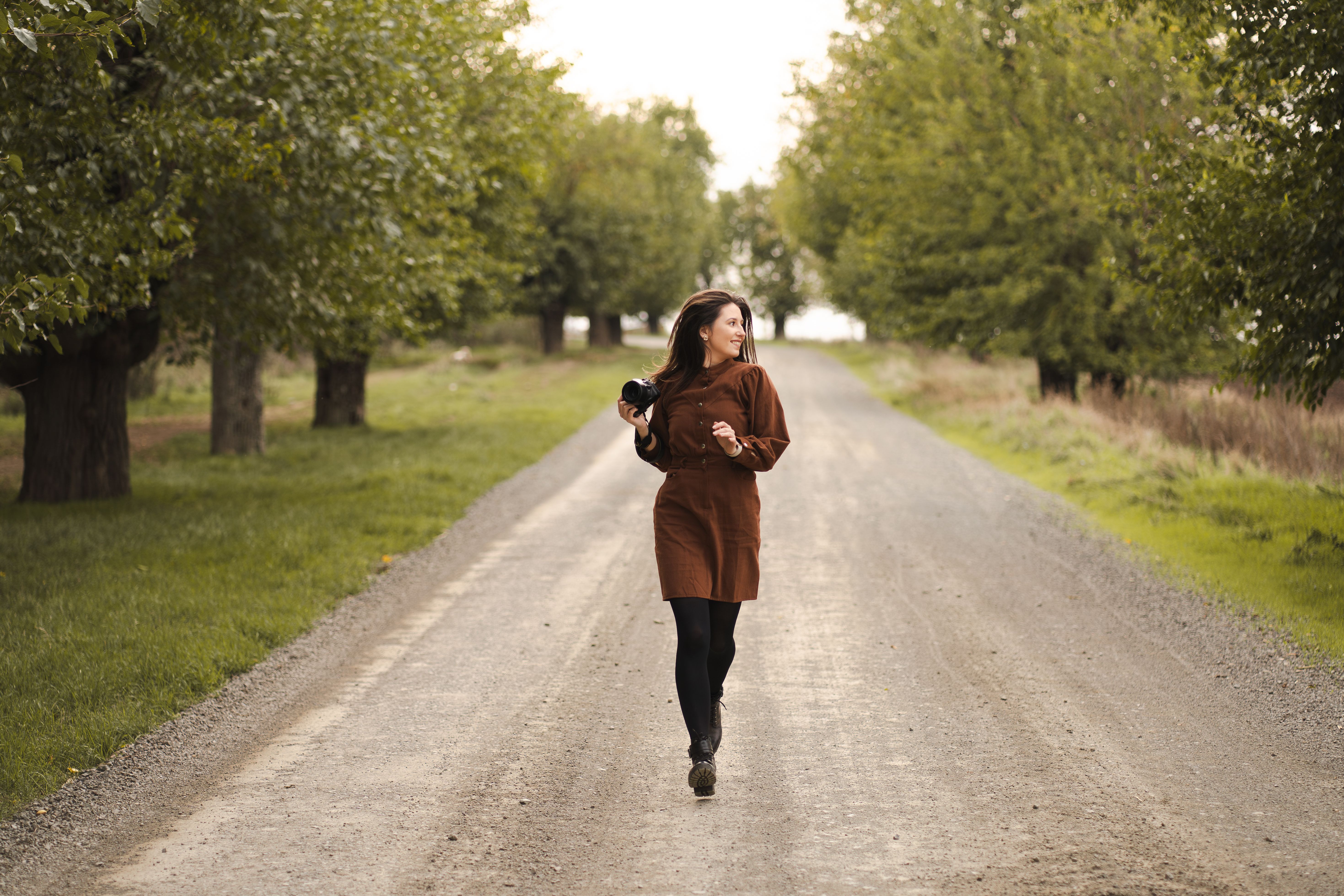 A woman photographer joyfully walks down a scenic tree-lined path in autumn.