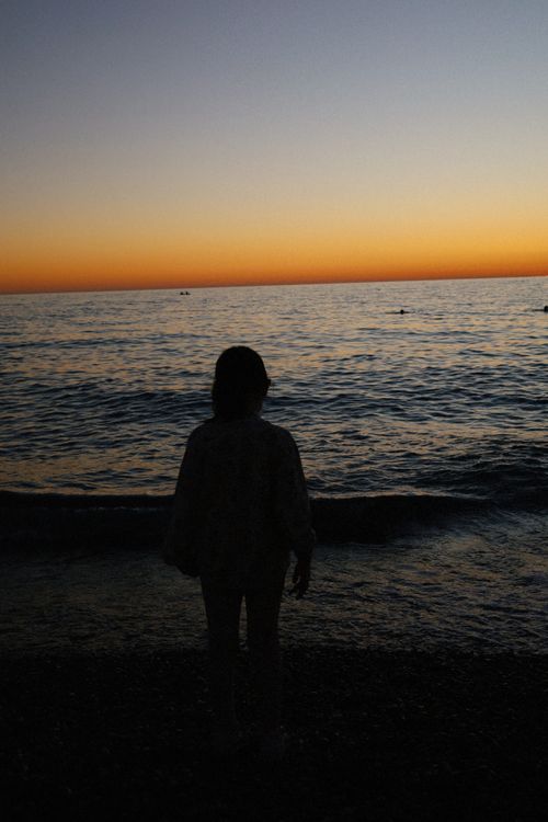 Free A person stands in silhouette facing the sea as the sun sets over the horizon. Stock Photo