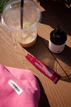 Skincare products and iced coffee on wooden table in sunlight.