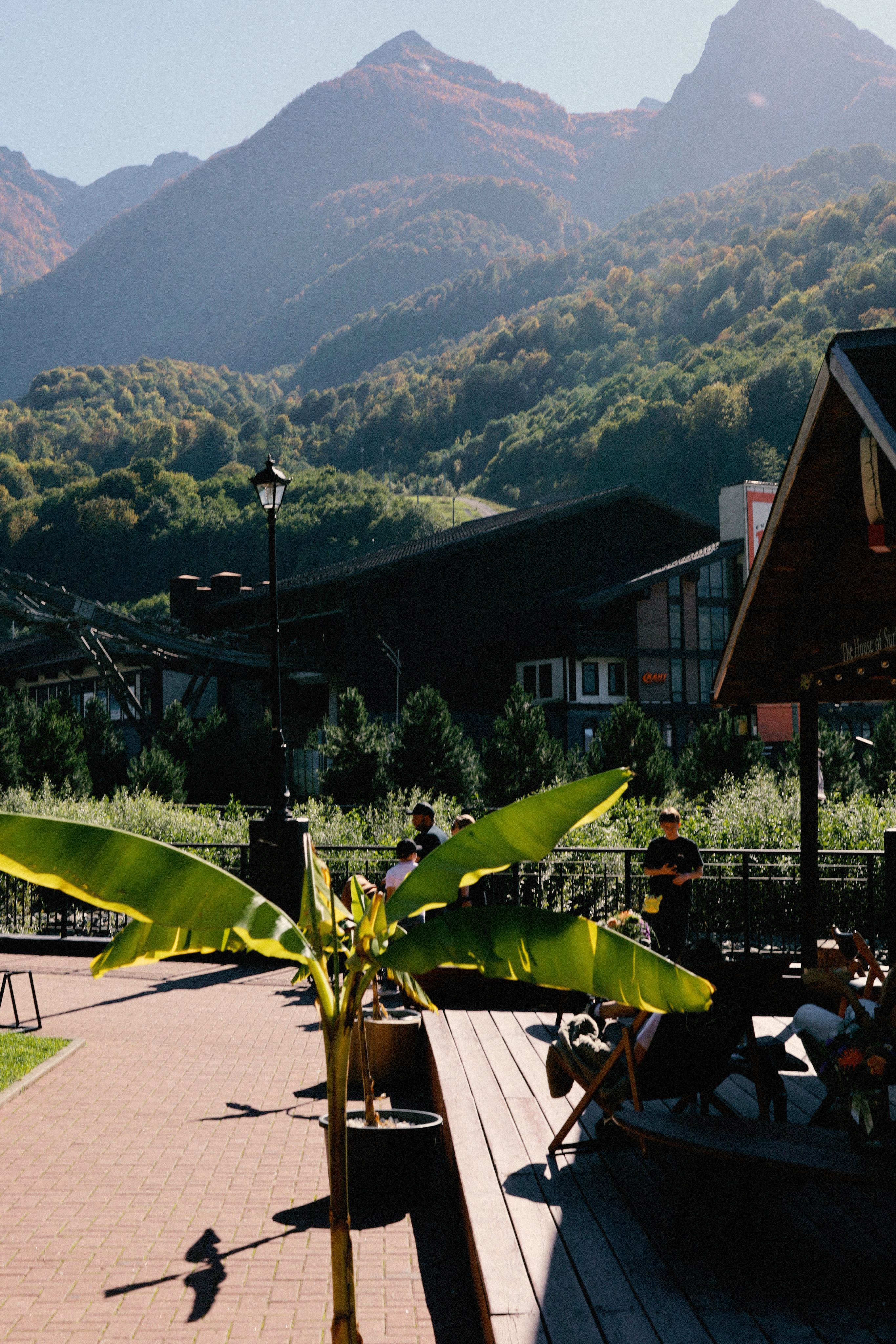 Scenic mountain view from a terrace with greenery, people, and sunny weather.