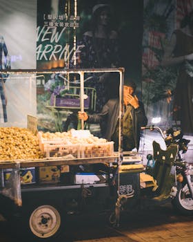 Street vendor at night market selling food on a motorized cart with bustling urban backdrop.