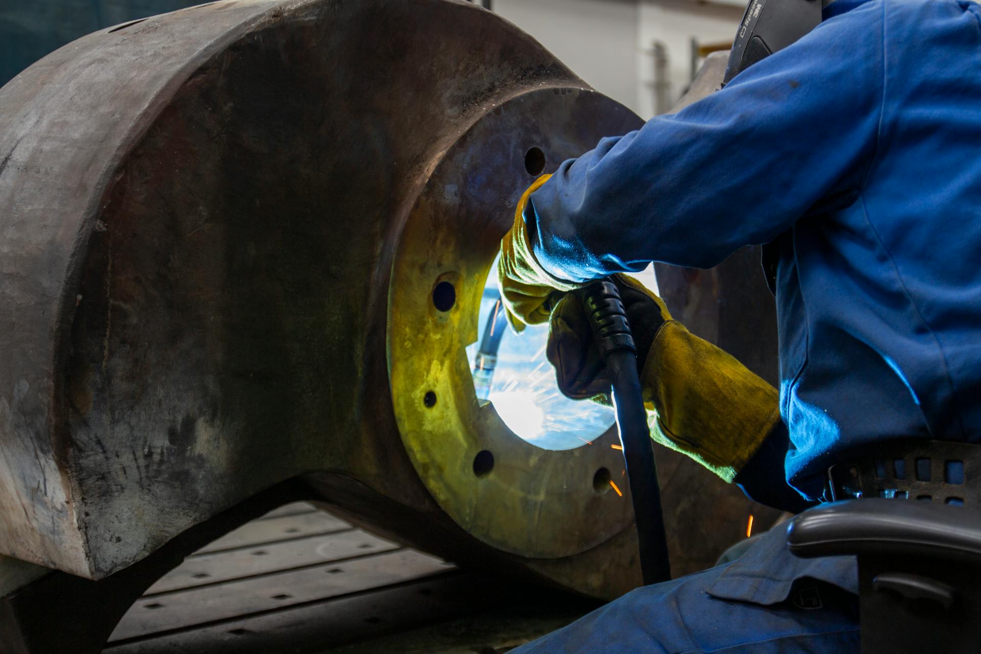 Welder operates inside a large metal machine part. Industrial setting with sparks visible.