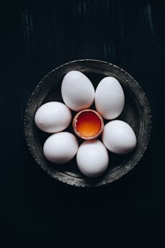 A dark, artistic arrangement of white eggs with one cracked to reveal a yolk.
