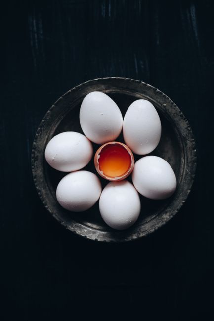 A dark, artistic arrangement of white eggs with one cracked to reveal a yolk.