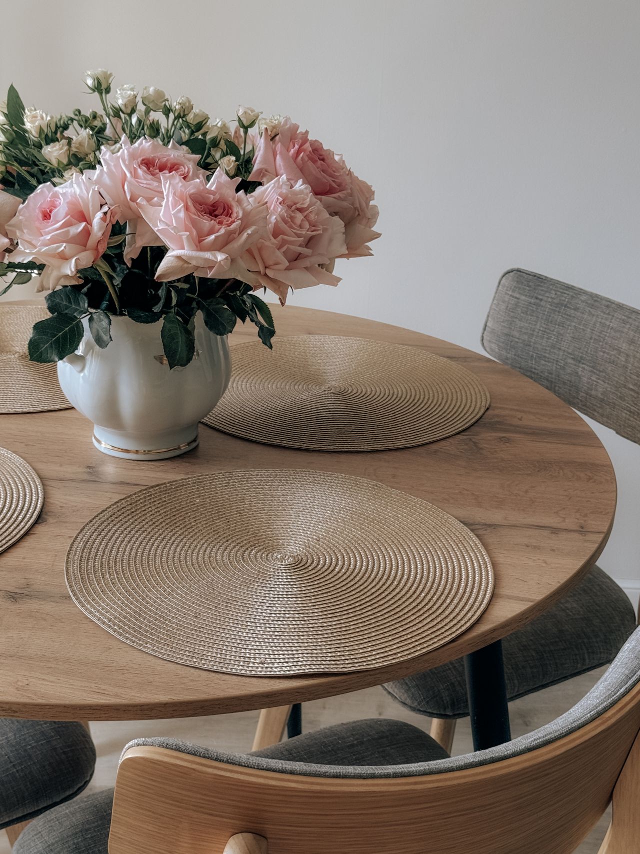 Round light wood table with woven placemats and a vase of pink roses