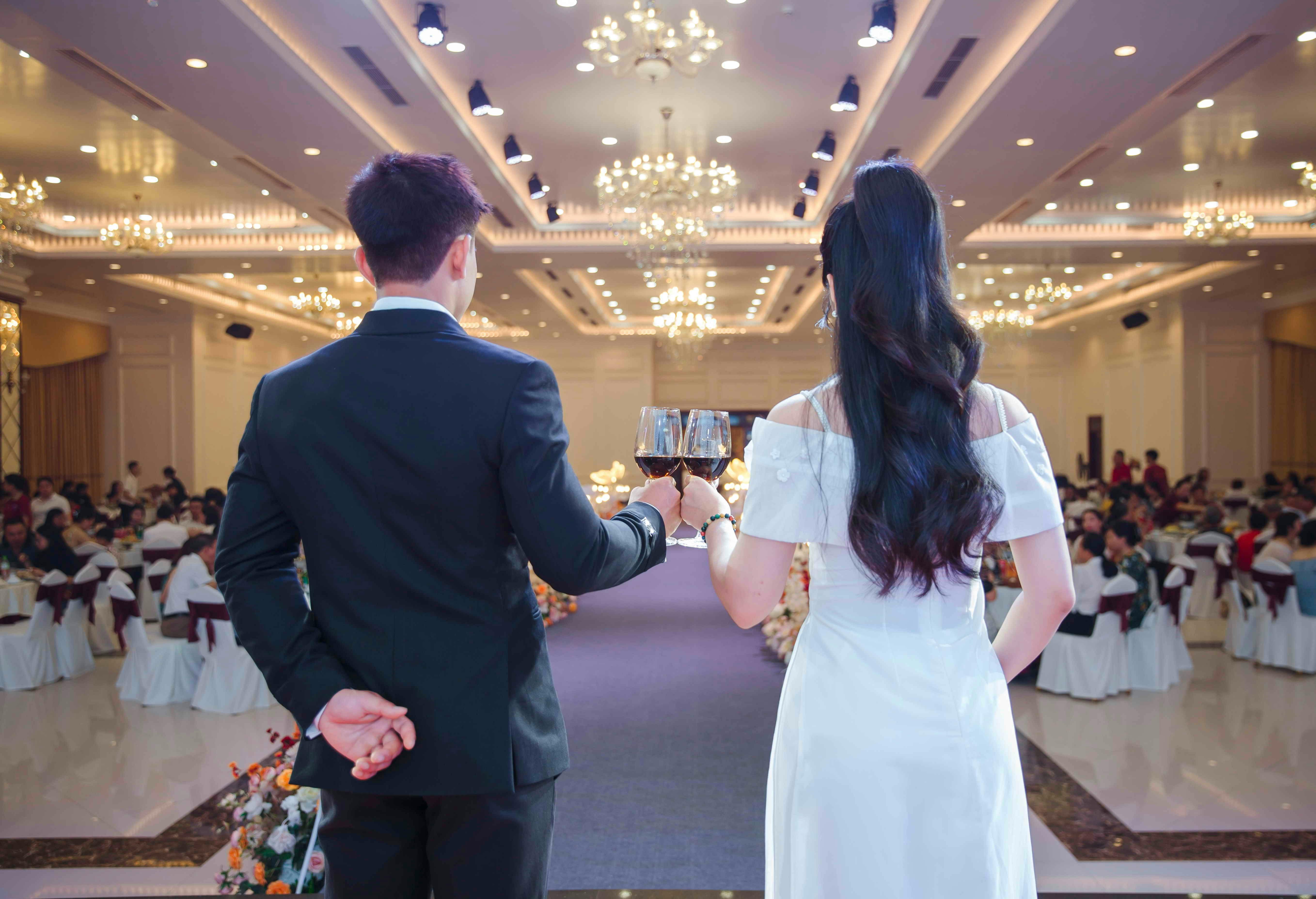 Back view of a couple toasting at a grand wedding reception in a lavishly decorated ballroom.