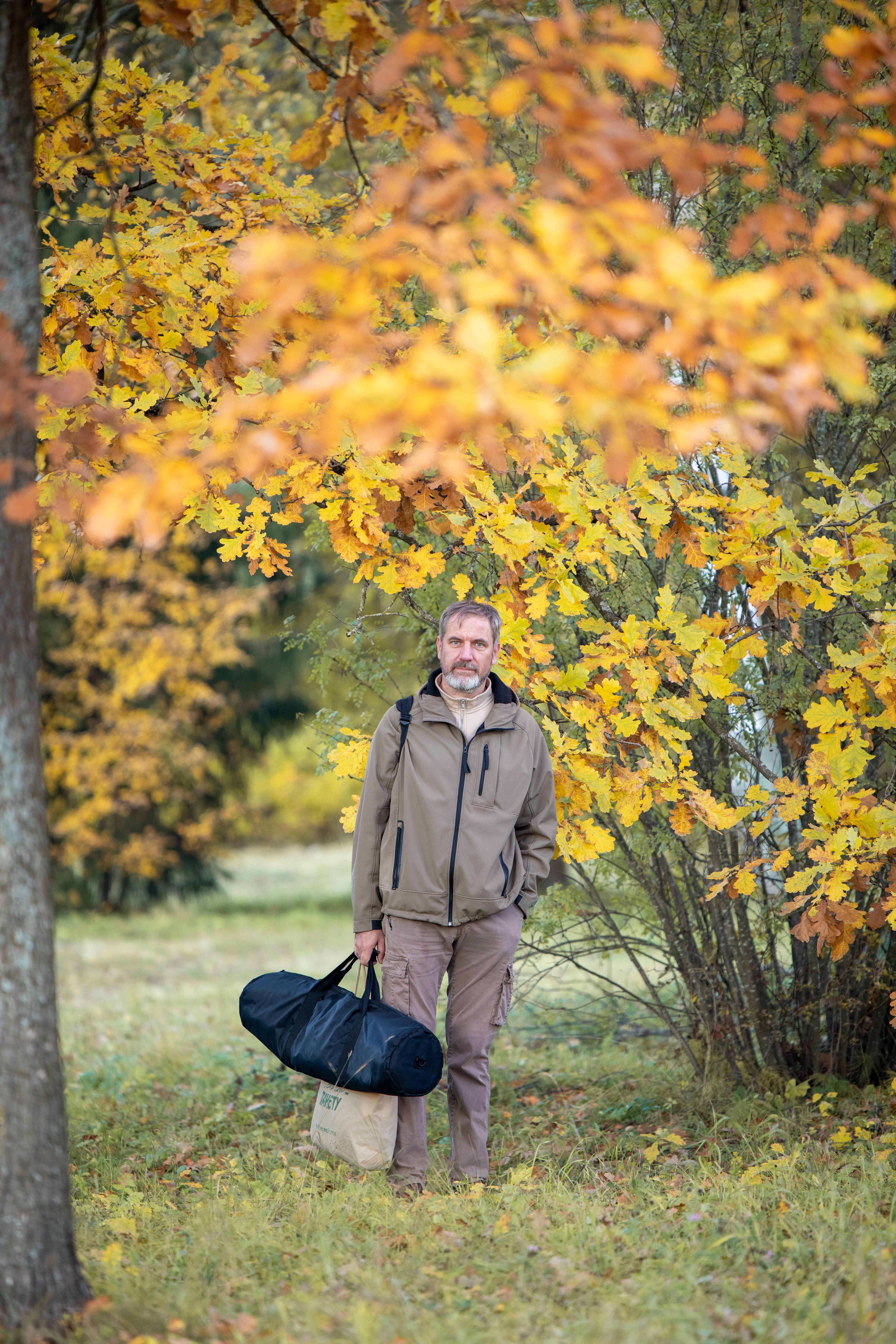 A man in a forest during autumn, holding a gear bag and surrounded by vibrant yellow foliage.