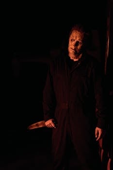 A mysterious masked figure stands with a knife in a dimly lit, eerie environment.