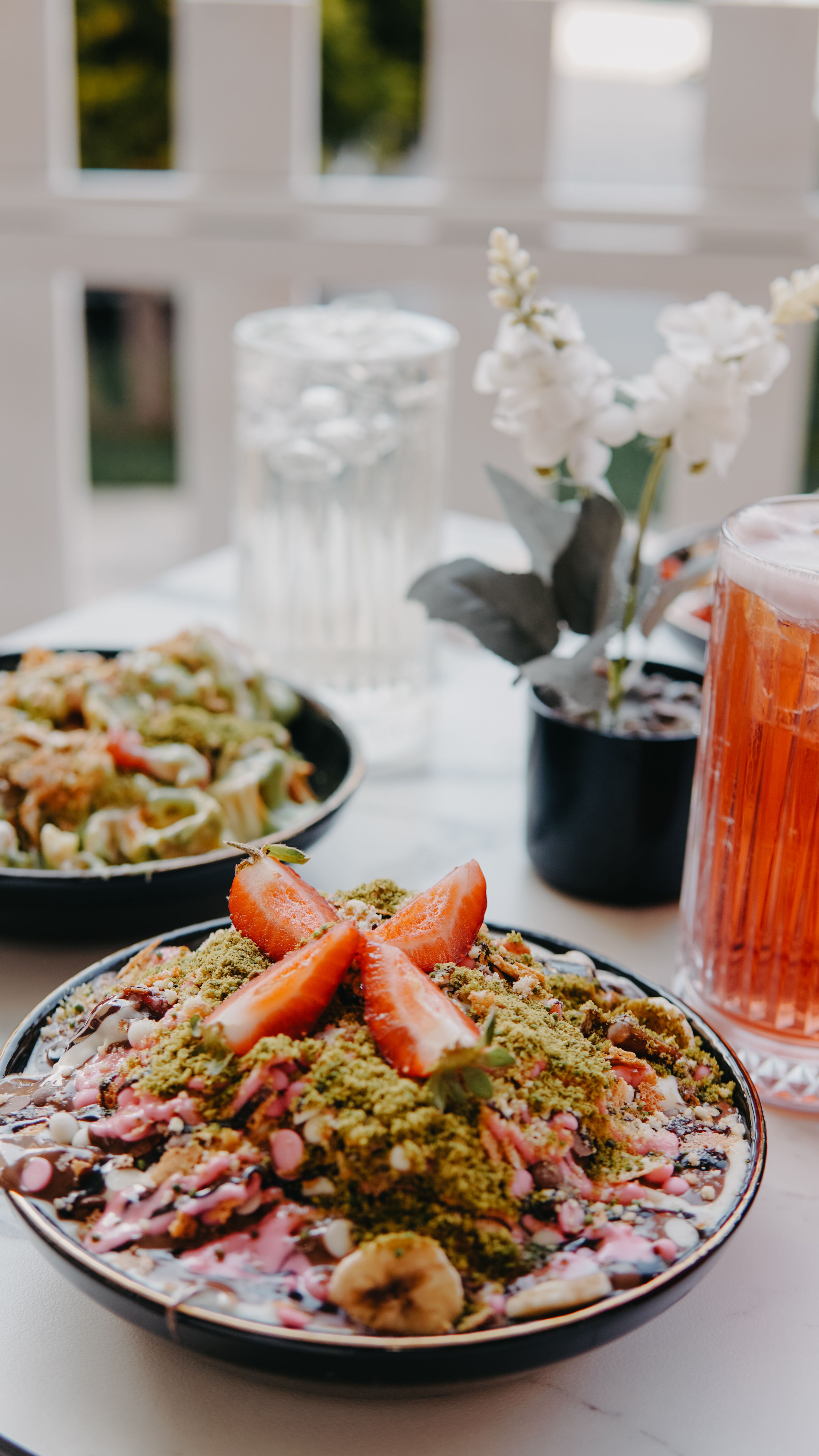 Strawberry and granola breakfast bowl with refreshing drinks and flowers on a table.