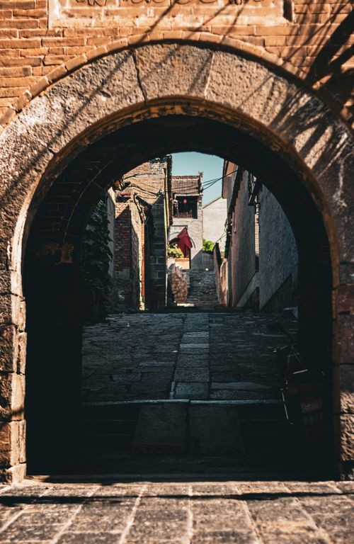 Free A historic stone archway framing an alley leading to sunlit traditional buildings. Stock Photo