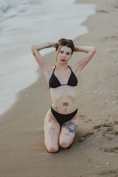 Woman kneeling on sandy beach in black bikini with tattoos visible, serene setting.