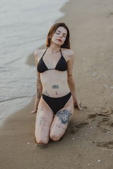 Woman with tattoos kneeling on sandy beach in black bikini, tranquil summer vibe.