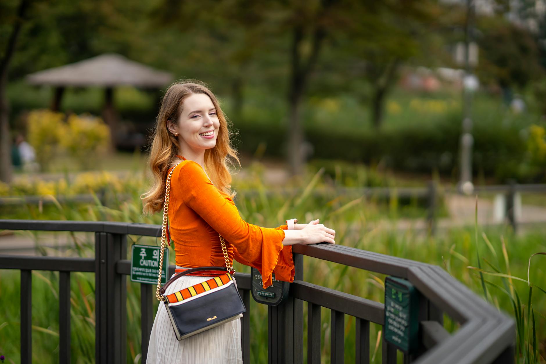 Woman in a bright orange top smiles on a park's wooden railing.