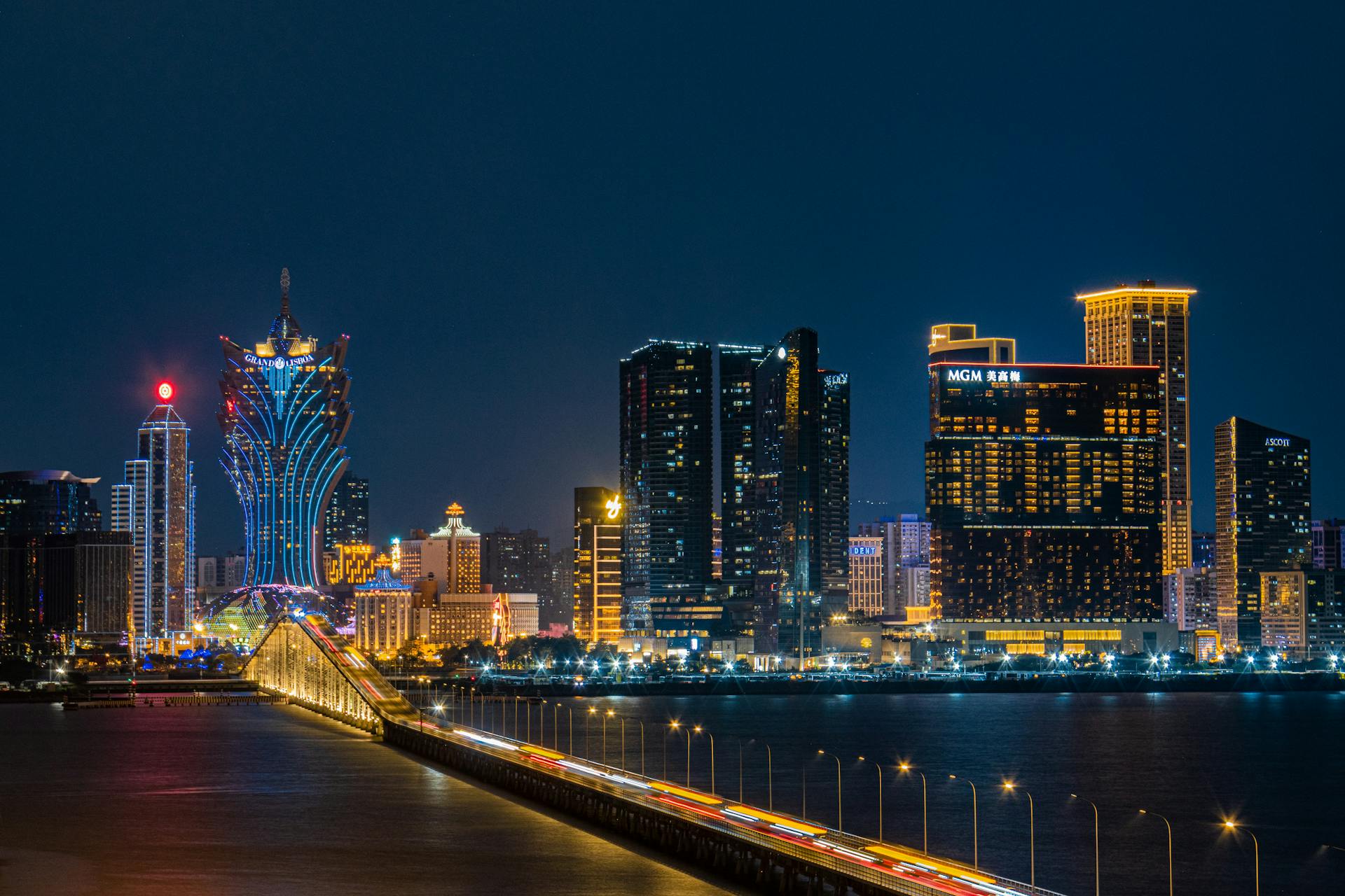 Elegant night view of Macau skyline featuring iconic illuminated hotels and a lit bridge over water.