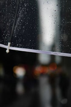 Transparent umbrella with raindrops focuses in a bustling city street with blurred lights.