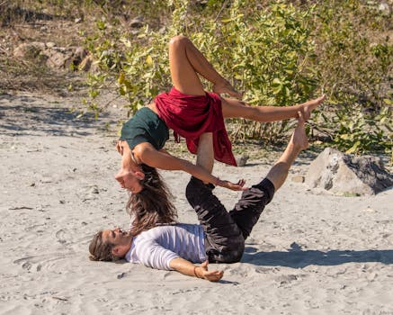 A couple performing an acro yoga pose on a sandy beach with natural background.