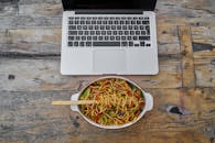 俯视质朴木桌，笔记本电脑与面条碗并置，展现食物与科技的完美融合