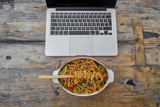 Overhead view of laptop and noodle bowl on rustic wooden table, perfect blend of food and technology.