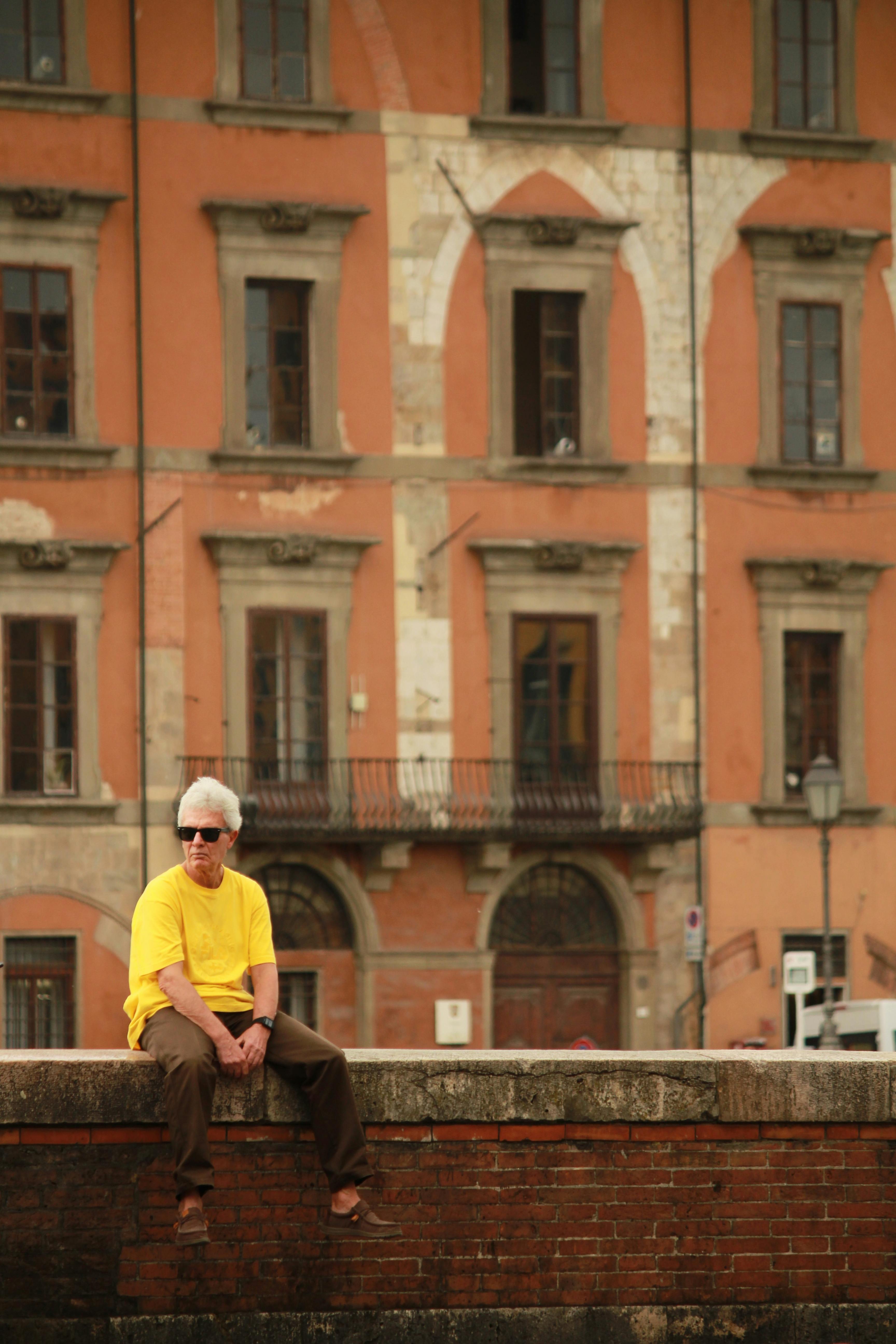 Senior man sits on wall in front of historic building in Pisa, Italy.