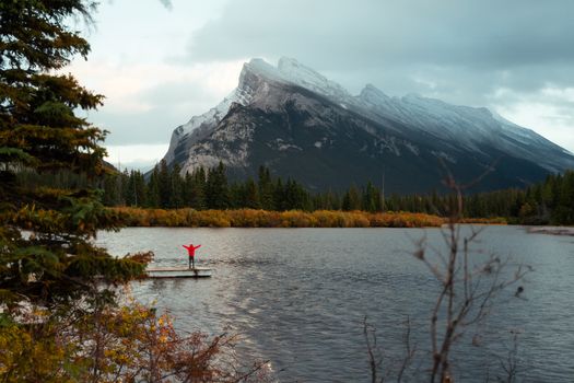 A lone figure in a red jacket stands by the lake with Mount Rundle in the background.