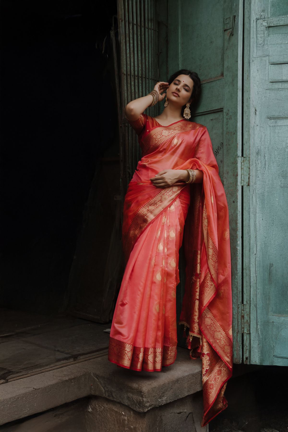 Model in a sindoor-red silk saree standing in a rustic doorway