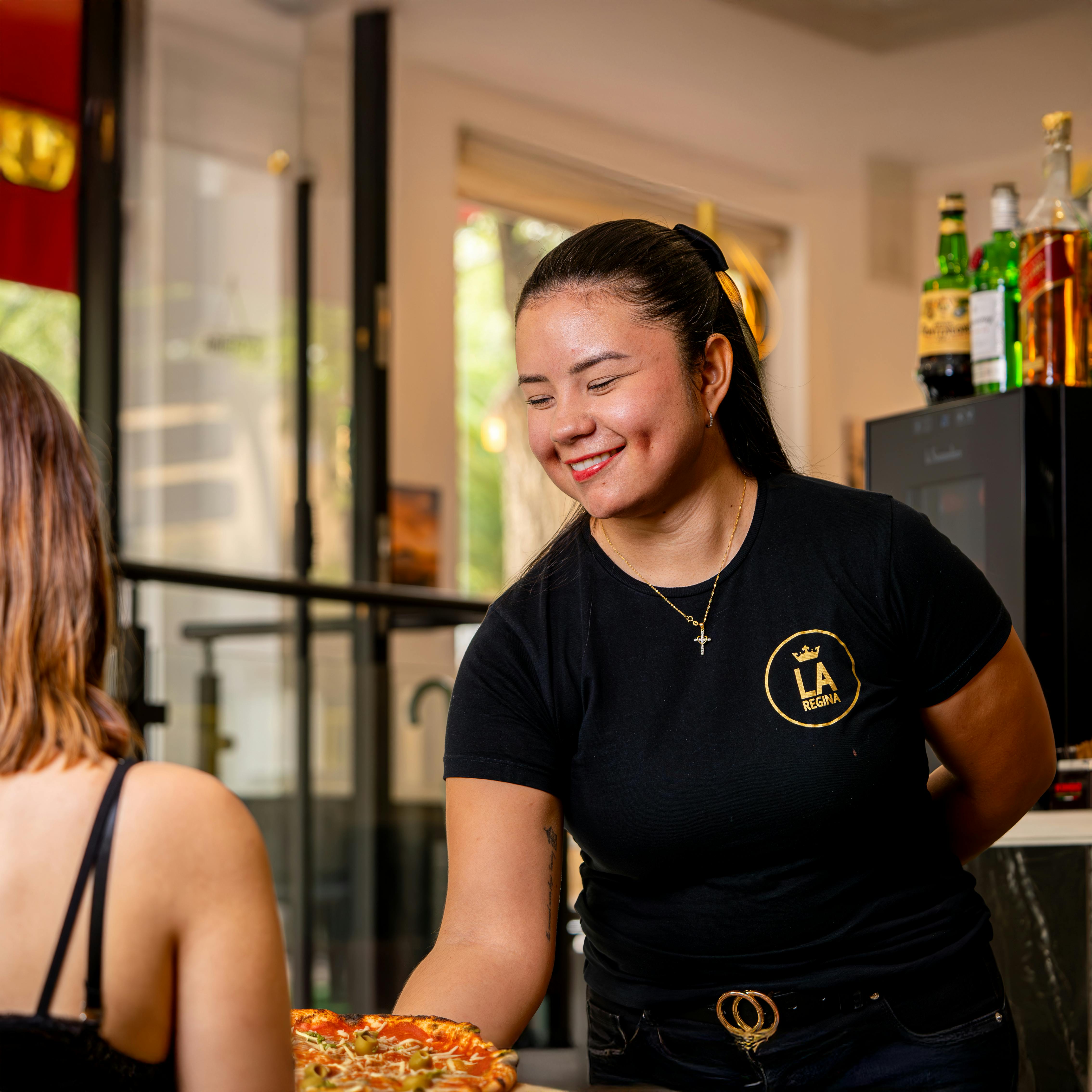 A friendly waitress serves a fresh pizza to a customer inside a cozy restaurant.