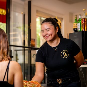 A friendly waitress serves a fresh pizza to a customer inside a cozy restaurant.