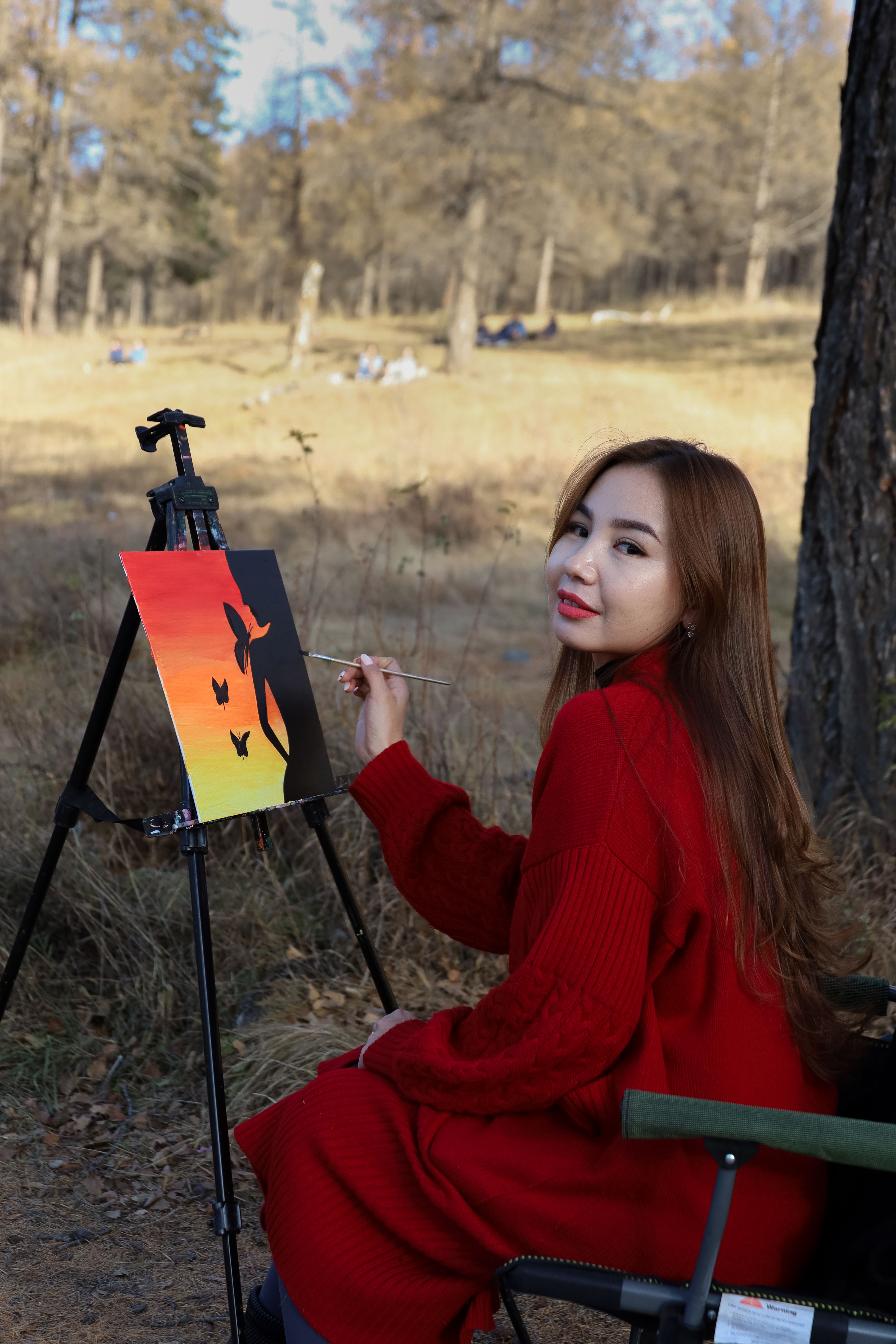 A woman in a red coat painting on a canvas outdoors in autumn.