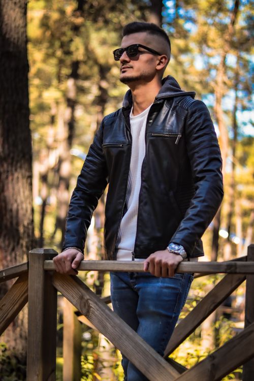 Free Young man wearing a leather jacket posing in a wooded park setting, highlighting casual fashion. Stock Photo
