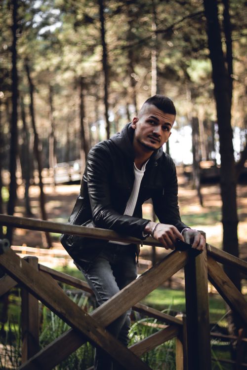Free Young man in casual attire leans on a wooden fence in a forest setting, exuding a relaxed vibe. Stock Photo
