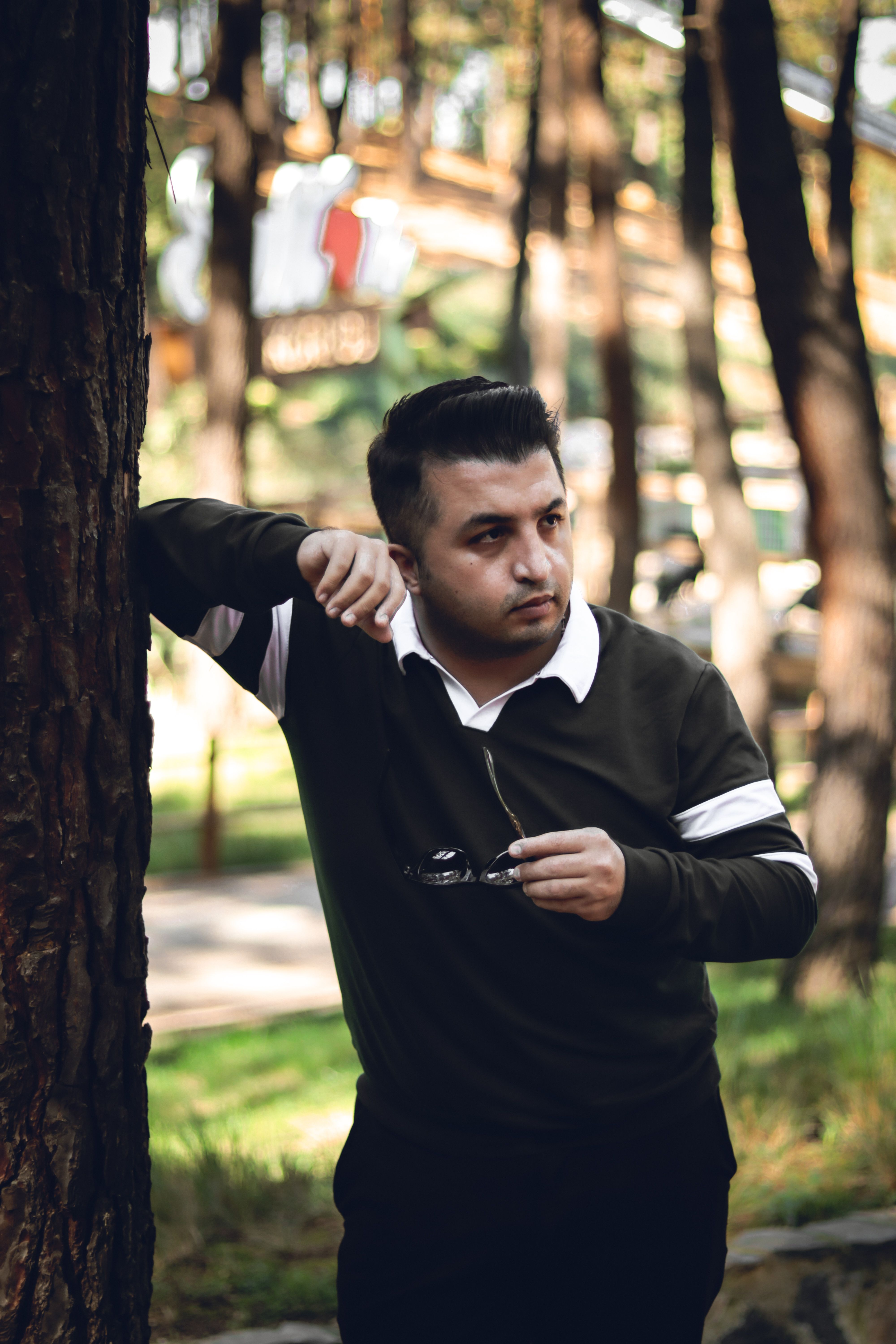 Man in casual attire leaning against tree in a sunny park setting, holding sunglasses.