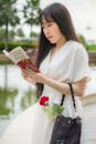 在越南胡志明市，一名身穿白色连衣裙的年轻女子手拿一朵红玫瑰，在湖边看书