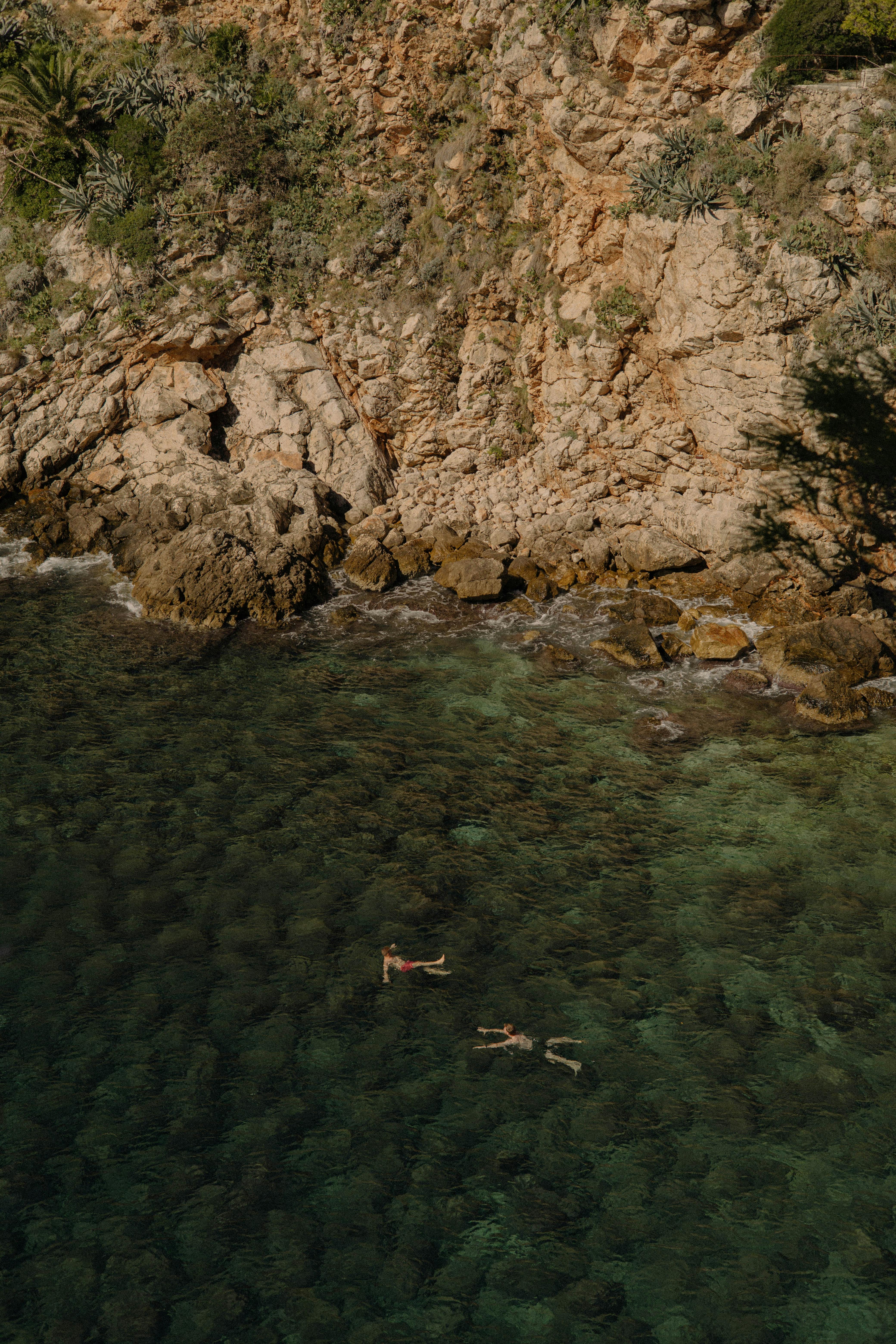 Aerial view of two swimmers exploring the clear waters by a rocky coastline.