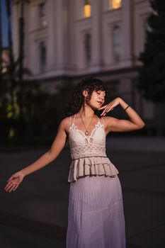 A woman in a stylish outfit poses gracefully outdoors against an elegant building at twilight.