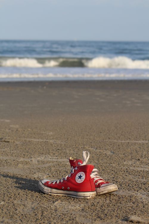 Free Red Converse sneakers laying on a sandy beach with ocean waves in the background, capturing a summer vibe. Stock Photo