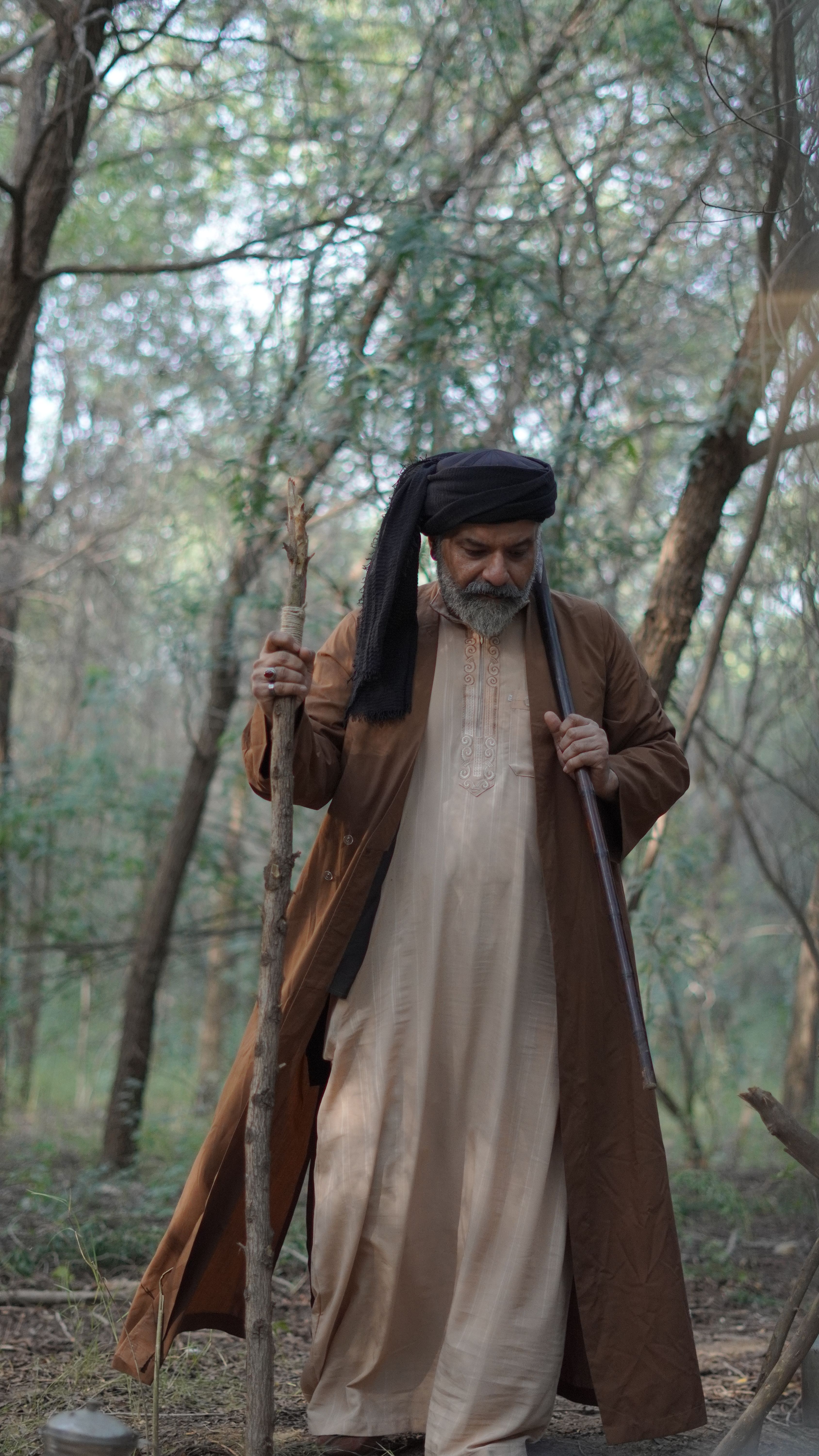 Elderly man in traditional attire walking with staff through a serene forest setting.