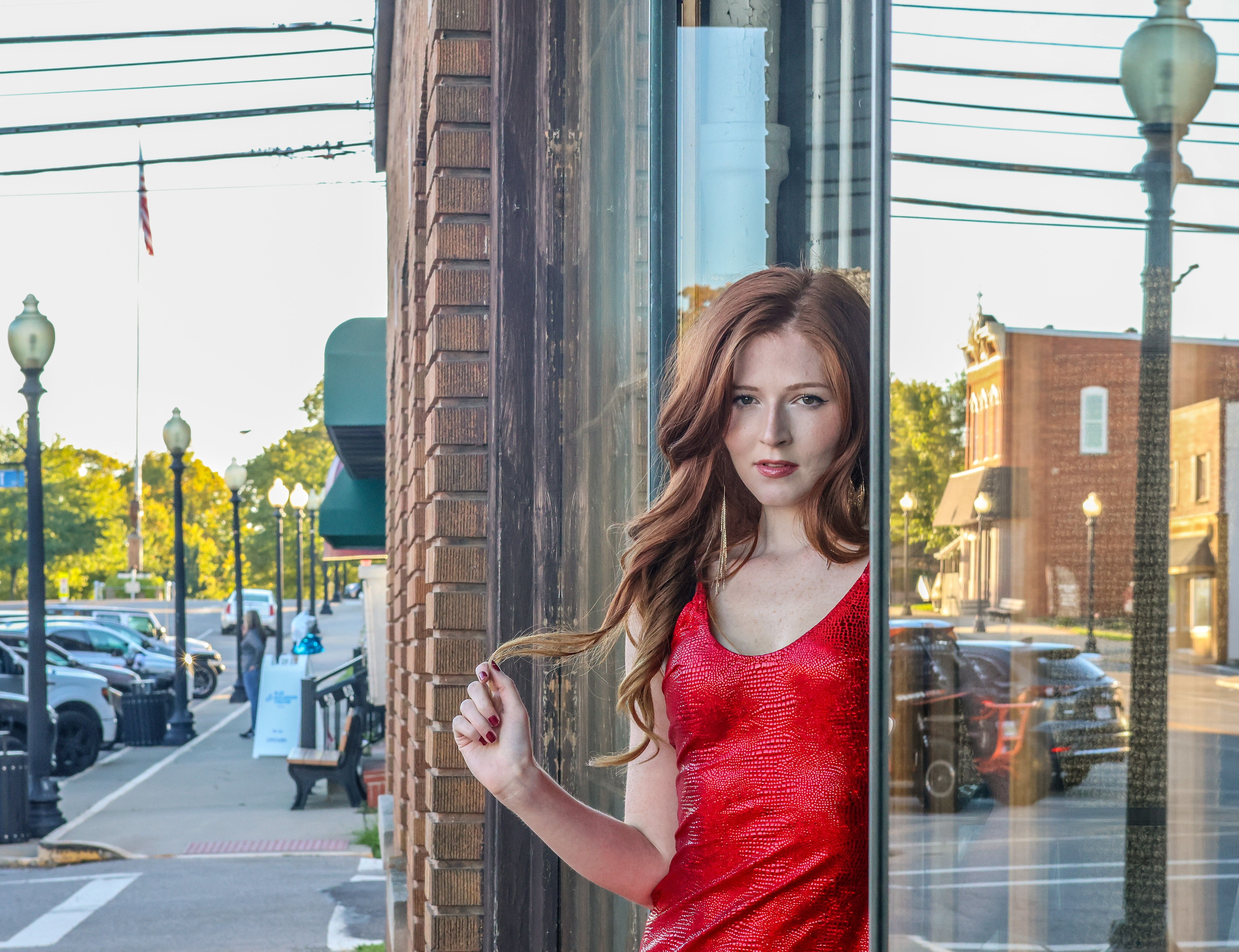 Elegant woman in red dress gazes through window in a small town setting.