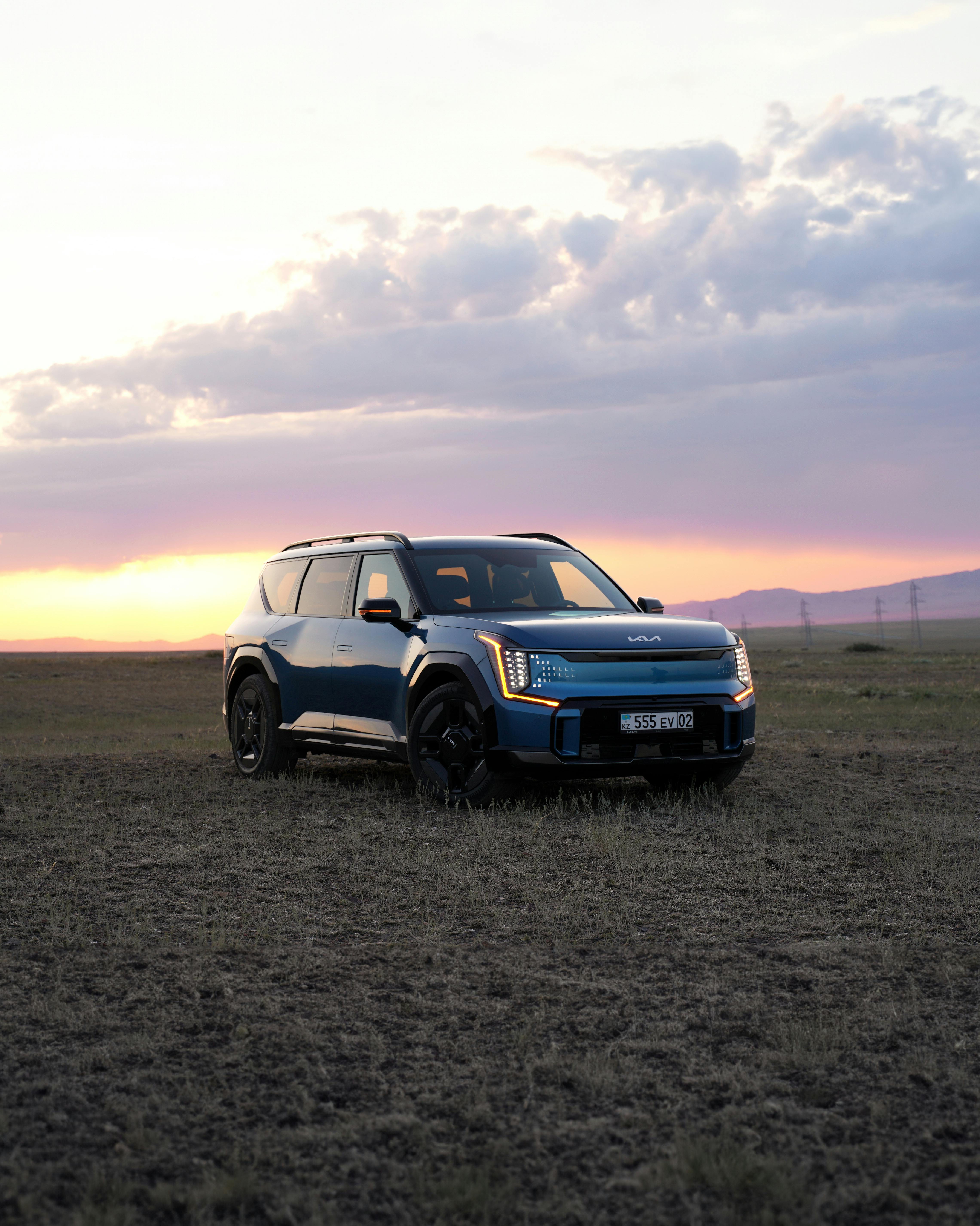 A blue electric SUV parked in a grassy field during sunset with dramatic sky and canyon backdrop.