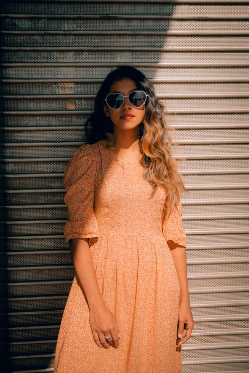 Free Stylish woman in peach dress and heart sunglasses posing against a shutter in bright sunlight. Stock Photo