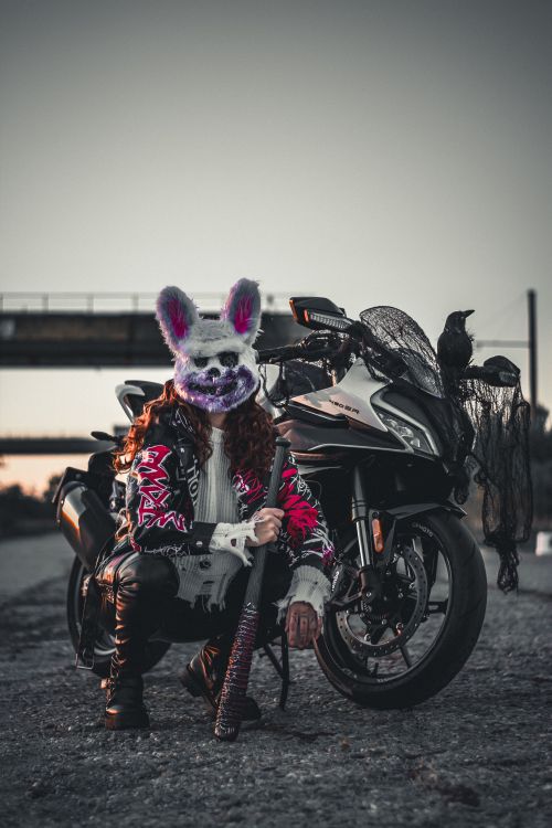 Free A striking image of a person in a bunny mask posing with a motorcycle outdoors, featuring a crow on the bike. Stock Photo