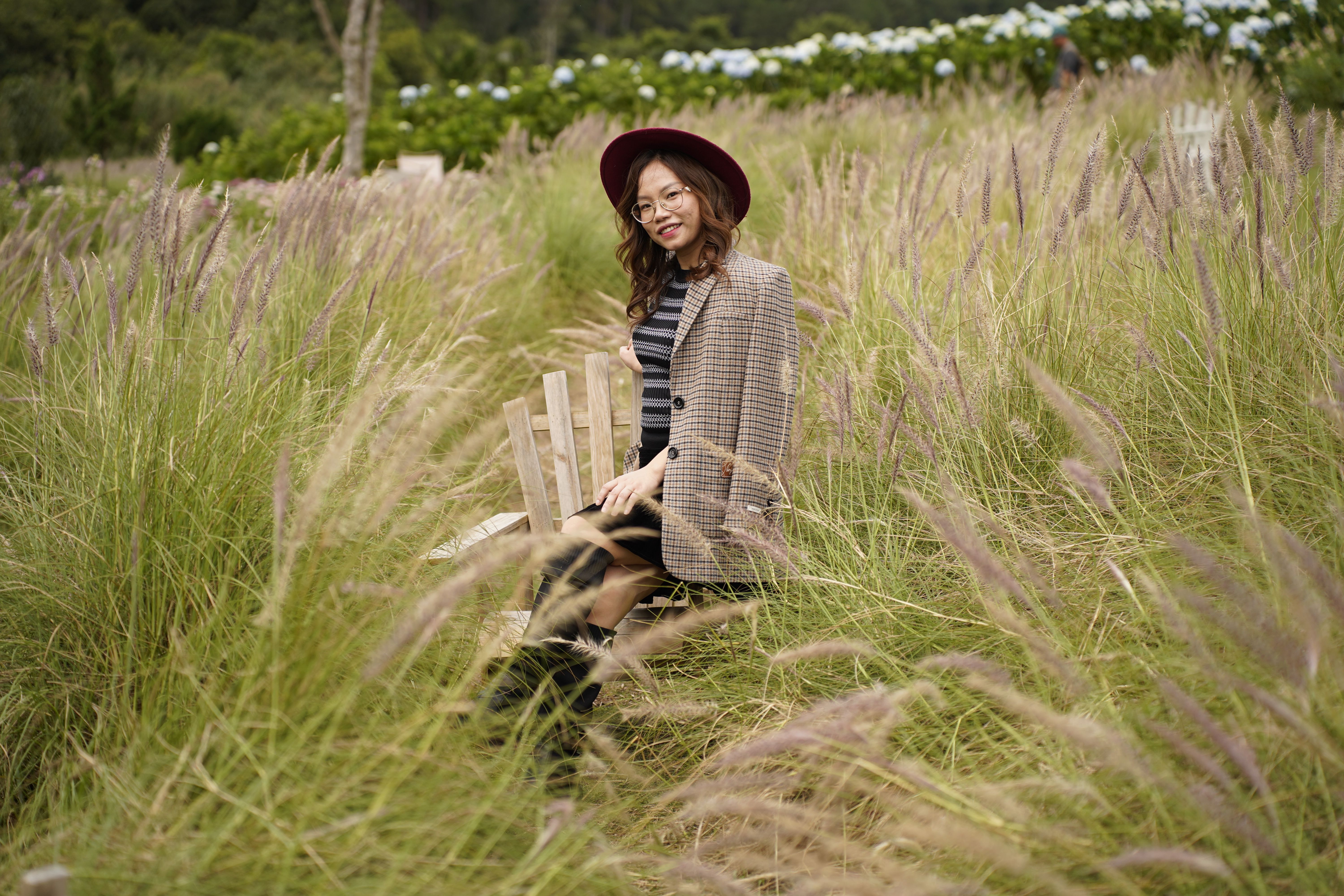 A woman in stylish attire sitting amidst a scenic meadow with tall grass and flowers.