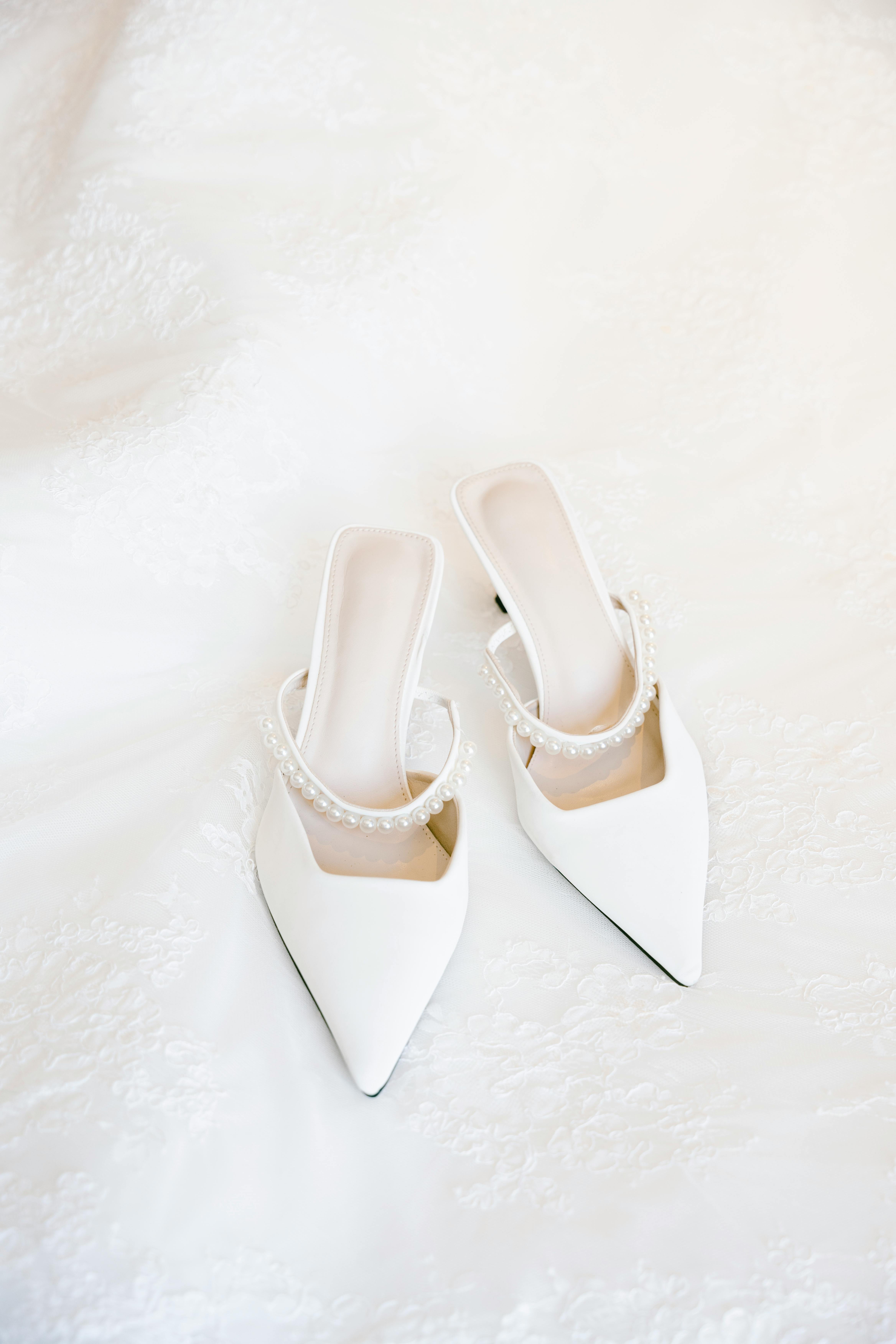 Stylish white pointed-toe bridal shoes with pearl details on a soft lace surface.